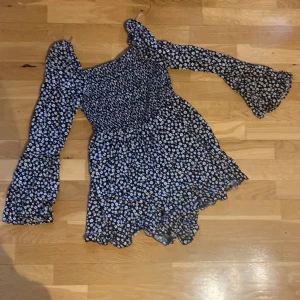 Blommig kortklänning från FB Sister XS - Supersöt svart kortklänning med vita blommor från FB Sister. Klänningen har långa trumpetärmar, smockad överdel och volangig nederkant. Perfekt för vår och sommar, med en luftig känsla och snygg passform. Shorts klänning 