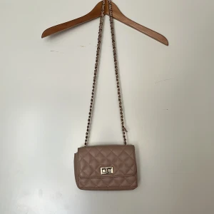 Beige quiltad axelväska med kedja - Säljer en beige/rosa quiltad axelväska med snygg guldfärgad kedjerem och vridlås. Lite sliten men väldigt snygg, men använder aldrig längre tyvärr 