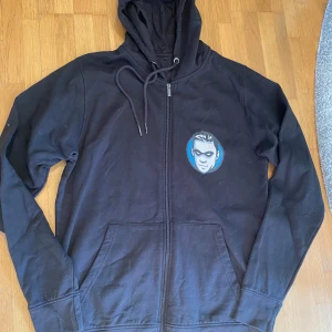 Svart hoodie med tryck och dragkedja - Svart hoodie med huva och snörning, hel dragkedja framtill och två fickor. På bröstet finns ett tryck