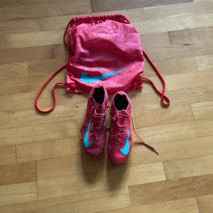 Rosa Nike fotbollsskor med väska ELITE - Säljer ett par snygga rosa Nike fotbollsskor med blå swoosh och matchande gympåse. Skorna har snörning och är tillverkade i syntetmaterial för bästa grepp och känsla på planen. Perfekta för dig som vill sticka ut på fotbollen!dem är storlek 43