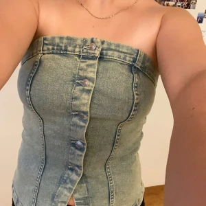 Ljusblå jeans tubtopp med knappar - Säljer en snygg ljusblå tubtopp i jeansmaterial med knappar framtill. Toppen är axelbandslös och har en tight passform som framhäver figuren. Perfekt till sommarens alla outfits och enkel att styla med både kjol och byxor.