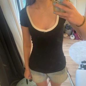 Mörkblå topp med vit kant och djup ringning - Säljer en mörkblå topp med korta ärmar och vit kant runt en djup, fyrkantig ringning. Modellen är tight och figurnära, perfekt för dig som gillar en enkel men snygg look. T-shirten är i mjukt material och passar till allt. Figursydd