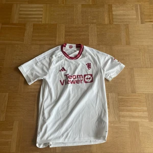Manchester United vit tredjetröja Adidas L - Säljer en vit Manchester United tredjetröja (säsong 23/24) från Adidas i storlek L. Tröjan har röda detaljer, klubbmärke och sponsortryck på bröstet samt röd-vit krage. Pris kan diskuteras!