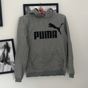 Grå hoodie från Puma med logga - Säljer en grå hoodie från Puma med stor svart logga och text på bröstet. Tröjan har huva, magficka och ribbade muddar vid ärmslut och nederkant. Perfekt för dig som gillar sportig och avslappnad stil.