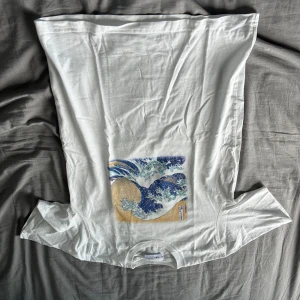 Vit t-shirt med vågtryck storlek S - Vit t-shirt från Uniqlo med ikoniskt vågmotiv i blått och beige framtill. Klassisk passform och rund hals. Tillverkad i mjuk bomull, perfekt för dig som gillar stilrena prints med japansk vibe.