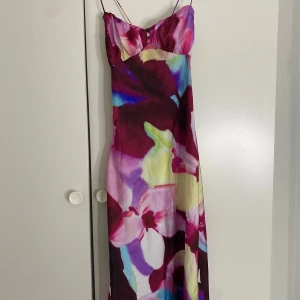 Lång blommig klänning med tunna band - Superfin långklänning med tunna axelband och färgstarkt blommigt mönster. Snygg v-ringning med små knappar framtill. Perfekt för sommarens alla tillfällen!