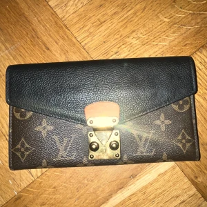 Louis Vuitton monogram plånbok - Snygg plånbok från Louis Vuitton i klassiskt monogrammönster. Framsidan har svart skinn och brunt LV-tryck, med guldfärgat spänne framtill. Perfekt storlek för kort och kontanter, och den har en lyxig känsla med sina detaljer i skinn och metall.