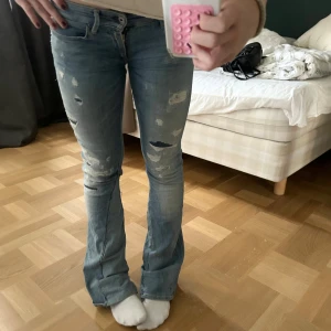 Lågmidjade slitna bootcut jeans - Säljer ett par blå jeans med slitningar och hål för en cool, avslappnad look. Modellen är bootcut med lätt utsvängda ben och klassiska fickor bak. Jeansen har låg midja och är gjorda i mjukt denimtyg som sitter snyggt på kroppen. Skriv för eventuella frågor💓