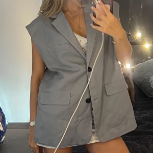 Oversized grå väst från Zara - Säljer en oversized grå väst från Zara med klassisk kavajkrage och två knappar framtill. Västen har två stora fickor med lock och en bröstficka. Perfekt att styla över en t-shirt eller klänning för en trendig look.