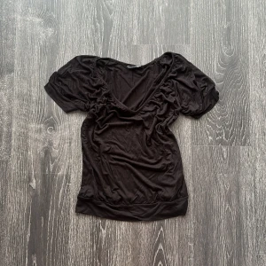 Svart v-ringad topp med puffärm - Svart topp med djup v-ringning och korta puffärmar. Modellen är figurnära med bredare kant nertill och lätt rynkade detaljer vid axlarna. Perfekt för dig som gillar en enkel men ändå trendig look.