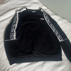 Svart sweatshirt från FILA med logga - Svart sweatshirt från FILA med vita detaljer och logoband längs ärmarna. Tröjan har rund halsringning, ribbade muddar och en broderad FILA-logga på bröstet. Perfekt för en sportig och avslappnad stil.