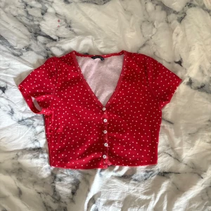 Röd croppad topp med hjärtan från SHEIN - Supergullig röd croppad topp från SHEIN med små vita hjärtan över hela. Toppen har korta ärmar, v-ringning och knappar framtill. Perfekt för dig som vill ha en söt och trendig look med lite 90-talsvibe.