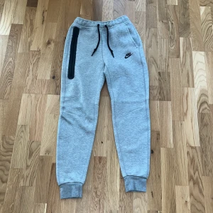 Grå Nike mjukisbyxor med dragkedja - Snygga ljusgrå mjukisbyxor från Nike med svart logga på låret och svarta snören i midjan. Byxorna har en lång svart dragkedja på ena benet och muddar vid bensluten för en skön passform. Perfekta för chill eller träning. Mjukisarna är i perfekt form har änvändst en gång men tyvärr va dom för små.