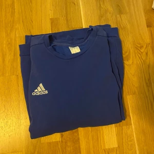 Blå Adidas sweatshirt 2XL - En klassisk blå sweatshirt från Adidas i storlek 2XL. Tröjan har rund hals, långa ärmar och den ikoniska Adidas-loggan broderad på bröstet samt tre vita ränder på ena ärmen. Perfekt för en sportig och avslappnad stil.