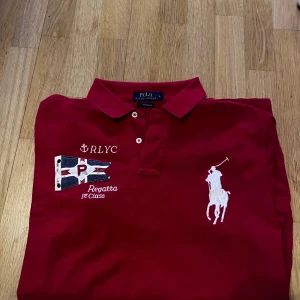 Röd pikétröja Polo Ralph Lauren - Snygg röd pikétröja från Polo Ralph Lauren med klassisk krage och knappar. Stor vit broderad logga på bröstet och coolt regatta-inspirerat tryck med flagga och text. Perfekt för en sportig och stilren look.