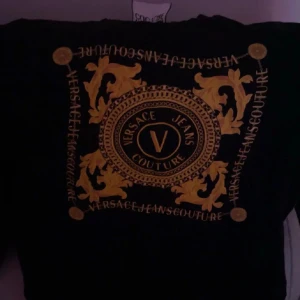 Svart t-shirt från Versace Jeans Couture - Svart t-shirt med stort guldigt tryck och dekorativa barockmönster på framsidan. Tydlig logga i mitten och runt om, klassisk rund hals och korta ärmar. Perfekt statement-plagg för dig som gillar lyxig stil.