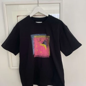 Svart t-shirt från maison margiela  - Svart t-shirt med ett stort färgglatt grafiskt tryck i rosa, gult och blått på bröstet. Klassisk passform och rund halsringning. Perfekt för dig som gillar unika prints och vill sticka ut lite extra.
