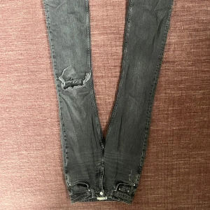 Svarta jeans med slitning - Säljer ett par svarta jeans med slitning på ena benet. Klassisk femficksmodell med dragkedja och knapp framtill. Jeansen har utsvängda ben och är i ett mjukt denimtyg.