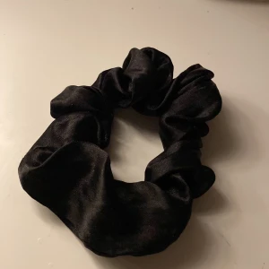 Svart scrunchie i satin - En klassisk svart scrunchie i glansigt satinmaterial. Perfekt för att sätta upp håret med en stilren och enkel look. Passar till många olika frisyrer och är skonsam mot håret.