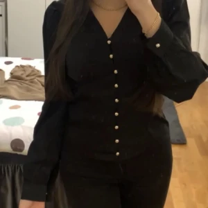 Svart blus med guldiga pärlknappar - Snygg svart blus med v-ringning och långa ärmar. Framsidan har guldiga pärlknappar hela vägen ner samt vid armar. Blusen har en feminin passform och lätt puff vid axlarna. Perfekt att styla med jeans eller kjol.