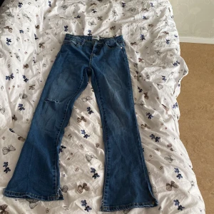 Blå bootcut jeans med rå kant - Säljer ett par blå bootcut jeans med rå, ofållad kant nertill. Jeansen har klassisk femficksdesign och en liten slitning på ena benet. Perfekta för dig som gillar en avslappnad och trendig look.