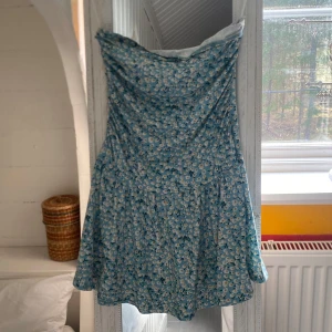 Blå blommig kortklänning - Supersöt blå klänning med vitt blommönster. Klänningen har en lätt utställd passform och är ärmlös, perfekt för varma dagar. Materialet känns lätt och luftigt, vilket gör den skön att bära under sommaren.