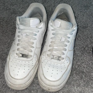 Vita Nike Air Force 1 sneakers - Klassiska vita Nike Air Force 1 sneakers med snörning och rund tå. Skorna har en platt sula och är helt vita med diskreta detaljer. Perfekta för en stilren och sportig look.