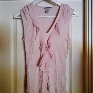 Ljusrosa ärmlös topp med volang från H&M - Säljer en ljusrosa ärmlös topp från H&M med v-ringning och dekorativa volanger framtill. Toppen är tillverkad i ett mjukt material av viskos och polyester, perfekt för vår och sommar. Använd enstaka gång, men den har ett litet lagat hål