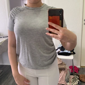 Grå t-shirt med rynkade axlar - Säljer en ljusgrå t-shirt med korta ärmar och snygga rynkade detaljer vid axlarna. T-shirten har en klassisk rund halsringning och är tillverkad i ett mjukt material som känns skönt mot huden.