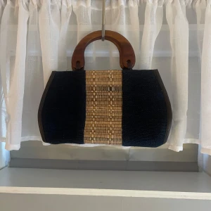 Svart och träfärgad handväska med trähandtag - Handväska med svarta sidor och mittparti i flätat rotting. Väskan har rundade trähandtag som ger en retro känsla.