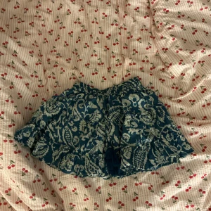 Blågrön kortkjol med blommigt mönster - Supersöt blågrön kortkjol med vitt blommigt mönster och volangdetaljer. Kjolen har en luftig känsla och passar perfekt för varma dagar. Snygg att matcha med en enkel topp eller stickad tröja.