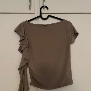 Beige topp med volangdetalj - Säljer en beige topp med asymmetrisk design och volang på ena axeln. Toppen har en kort ärm på ena sidan och volangdetaljer som ger en unik look. Perfekt för dig som vill sticka ut lite extra.