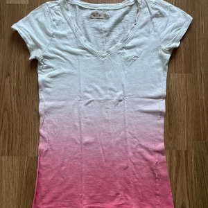 Vit och rosa t-shirt från Hollister - Vit till rosa ombre t-shirt från Hollister med v-ringning och korta ärmar. T-shirten har en liten broderad logga på bröstet och är gjord i mjuk bomull. Perfekt för en avslappnad stil.