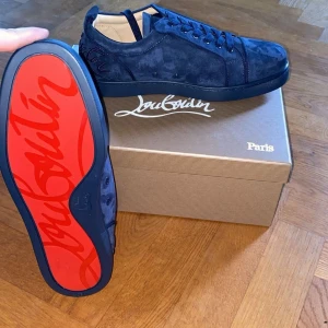 Marinblå mocka skor från Christian Louboutin - Säljer ett par exklusiva blå sneakers från Christian Louboutin i mocka med klassisk röd sula. Skorna har snörning och rund tå, och levereras med originalkartong, dustbags och skohorn. Perfekta för dig som vill sticka ut med stil.