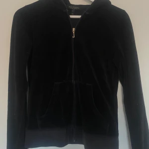 Svart velour hoodie från Juicy Couture - Säljer en svart velour hoodie från Juicy Couture med dragkedja och huva. Jackan har glittrig Juicy Couture-logga på ryggen och fickor framtill. Perfekt för en trendig och avslappnad look.