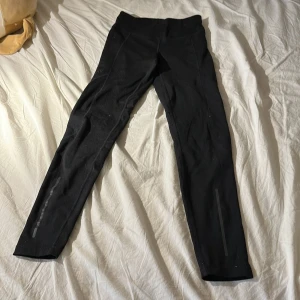 Svarta träningstights från Craft - Svarta tights från Craft i stretchigt syntetmaterial. De har en tight passform och reflekterande detaljer på benen samt snörning i midjan. Perfekta för träning och löpning.