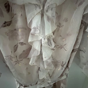 Ljusblommig volang jumpsuit med knytband - Superfin jumpsuit i ljus färg med blommigt mönster och subtila guldstjärnor. Jumpsuit har volangdetaljer längs v-ringningen och ärmarna, samt ett knytband i midjan. Perfekt för vår och sommar!