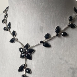 Blommigt halsband med svarta stenar - Elegant halsband med silverfärgad kedja och svarta, facetterade stenar formade som blommor och blad. Justerbar längd med kedja baktill. Perfekt för att ge en stilren touch till din outfit.