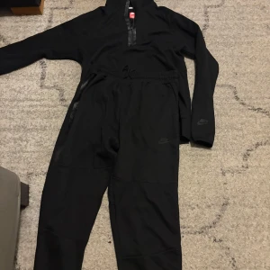 Svart träningsset från Nike - Säljer ett svart träningsset från Nike med långärmad tröja och matchande byxor. Tröjan har halv dragkedja framtill och en ficka med dragkedja. Byxorna har resår i midjan och dragkedjeförsedda fickor. Perfekt för träning eller sportiga dagar.