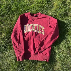 Vinröd sweatshirt från Dickies - Säljer en vinröd sweatshirt från Dickies med stor gul och vit logga framtill. Tröjan har rund halsringning, långa ärmar och ribbade muddar vid ärmslut och nederkant. Perfekt för en avslappnad stil.