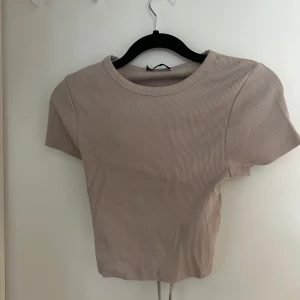 Beige ribbad croppad topp med snörning i ryggen - Säljer en beige ribbad croppad topp med korta ärmar. Toppen har en snygg snörning i ryggen som ger en unik look. Perfekt att styla till jeans eller kjol för en trendig outfit.