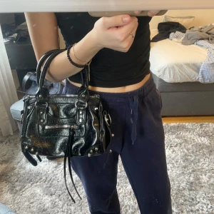 Svart handväska  - Säljer en svart handväska  i glansigt skinn med silverfärgade metalldetaljer och dragkedja framtill. Väskan har dubbla handtag och dekorativa remmar. Perfekt för att lyfta vilken outfit som helst.