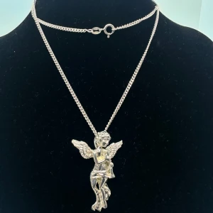 Halsband med ängelberlock i 925 äkta silver  - Snyggt halsband med en detaljrik ängelberlock i 925 silver. Kedjan är tunn och har ett klassiskt lås. Perfekt för dig som gillar unika smycken med lite edge och symbolik. Kedja och hänge i 925 äkta silver. Längd: 59,5cm, hänge storlek: ca 37x27mm. Kedjan väger 5,9g, 2mm. 