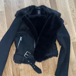 Svart jacka med fuskpäls och bälte  - Snygg svart jacka  med mjuk fuskpäls på kragen och insidan. Jackan har dragkedjor, bälte i midjan och långa ärmar. Perfekt för kyligare dagar och ger en cool look.