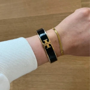 Svart och guld armband från Tory Burch - Snyggt armband från Tory Burch i svart och guld med det ikoniska logomärket framtill. Armbandet har en stilren design med blank finish. Lite skavt (se bilder).  Nypris 2000kr. 