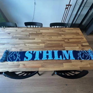 Djurgården halsduk med motivet ”STHLM” på. - Säljer en lång halsduk i mörkblått och ljusblått med STLM-tryck och grafiskt ansiktsmönster på sidorna. Halsduken har fransar i ändarna och är perfekt för att visa ditt stöd på Djurgårds matcher perfekt!