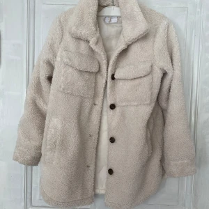Beige teddyjacka från Lindex - Mysig beige teddyjacka från Lindex med stora bröstfickor och knäppning framtill med bruna knappar. Jackan har lång ärm och är tillverkad i ett mjukt, fluffigt material som passar perfekt för kyligare dagar.
