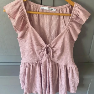 Rosa volangtopp från 157 - Söt ljusrosa topp från 157 med volangärmar och knytdetalj framtill. Toppen har en lätt rynkad struktur och v-ringning, samt en lös passform nedtill. Perfekt för vår och sommar!