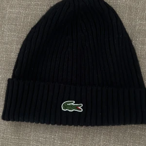 Svart ribbad mössa från Lacoste - Svart ribbstickad mössa från Lacoste med klassisk krokodillogga framtill. Tillverkad i 100% ull för extra värme och komfort. Perfekt för kyliga dagar och enkel att matcha med olika outfits.