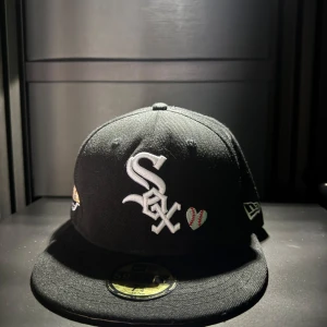 Svart keps från New Era x White Sox - Svart keps med White Sox-logga framtill, broderade detaljer som ett hjärta med baseball-sömmar, texten 'Love & Hustle 4 the Game' bak och World Series-märke på sidan. Klassisk rak skärm och broderad New Era-logga.